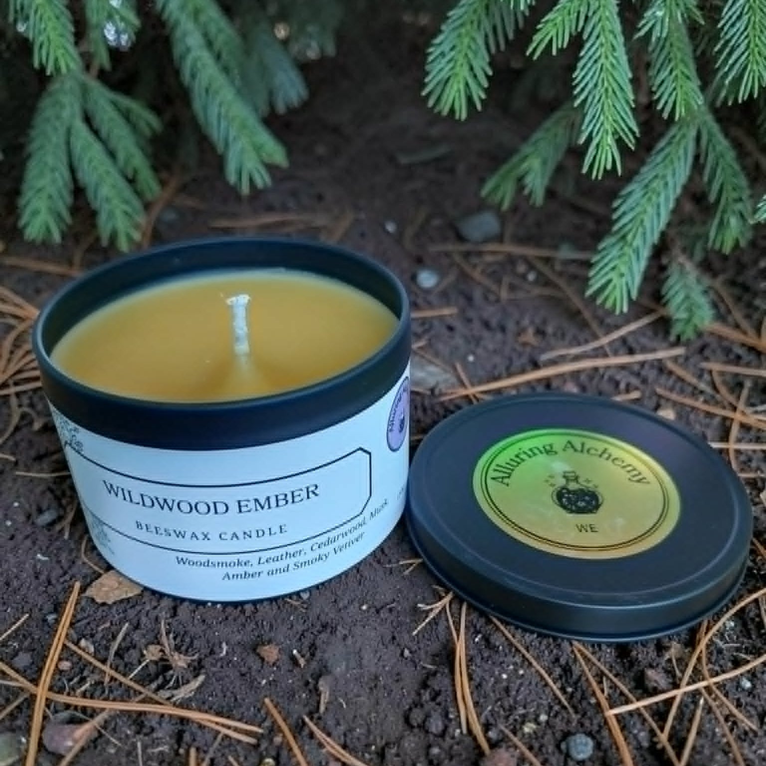 Candle in a tin labeled 'Wildwood Ember' on the ground with green foliage in the background