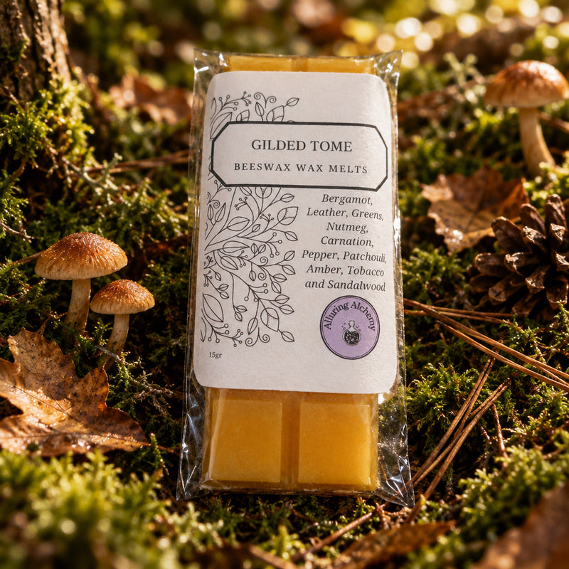 A packaged bar of beeswax melt labeled 'Gilded Tome' by The Queeny Academy, showing two yellow wax bars in clear plastic packaging on a light wood surface.