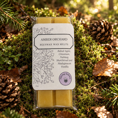A packaged stick of Amber Orchard Beeswax Wax Melts on a forest floor. The wax melt stick is visible through clear plastic packaging with a white label featuring black text and botanical line drawings.