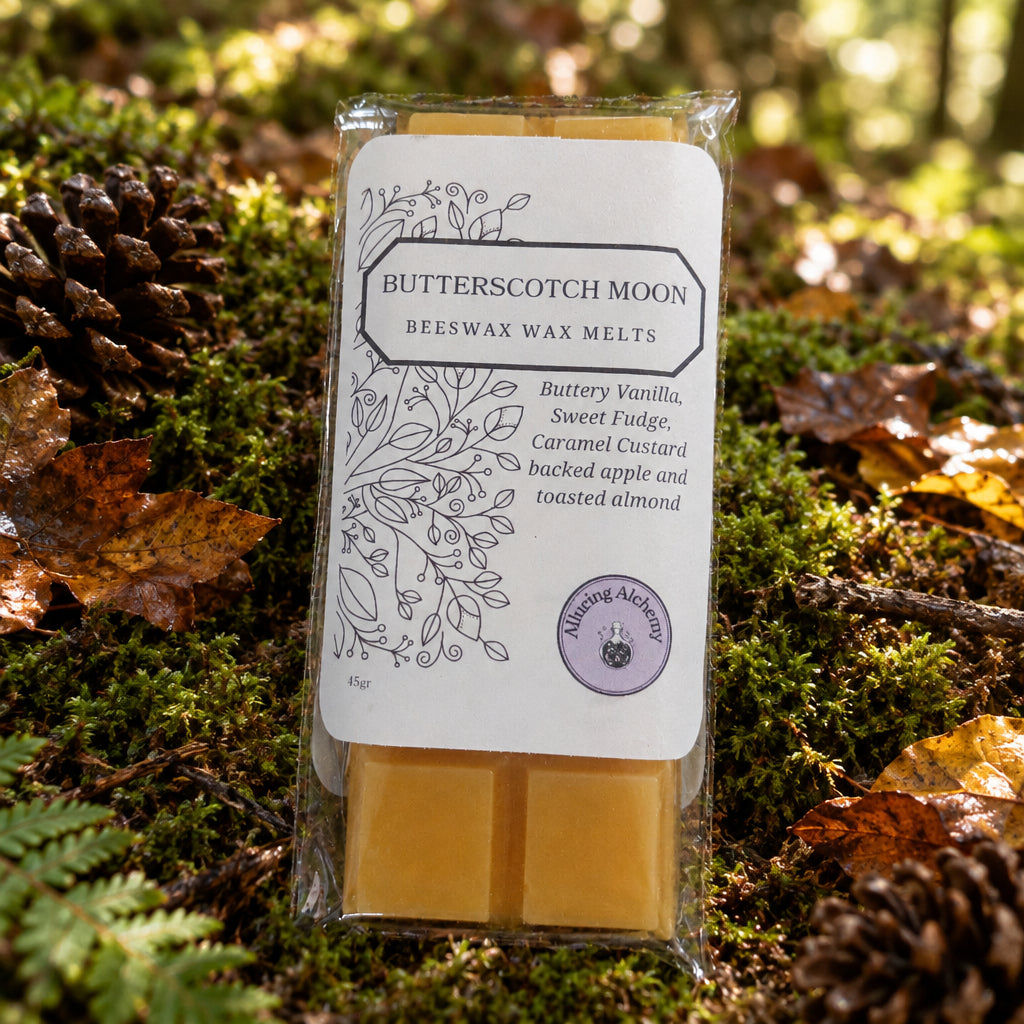 A packaged bar of beeswax wax melts labeled 'Butterscotch Moon' sits on a light wood surface. The clear plastic wrapper holds two pale golden wax bars with a white paper label featuring black text and botanical line art, plus a small purple circular logo.