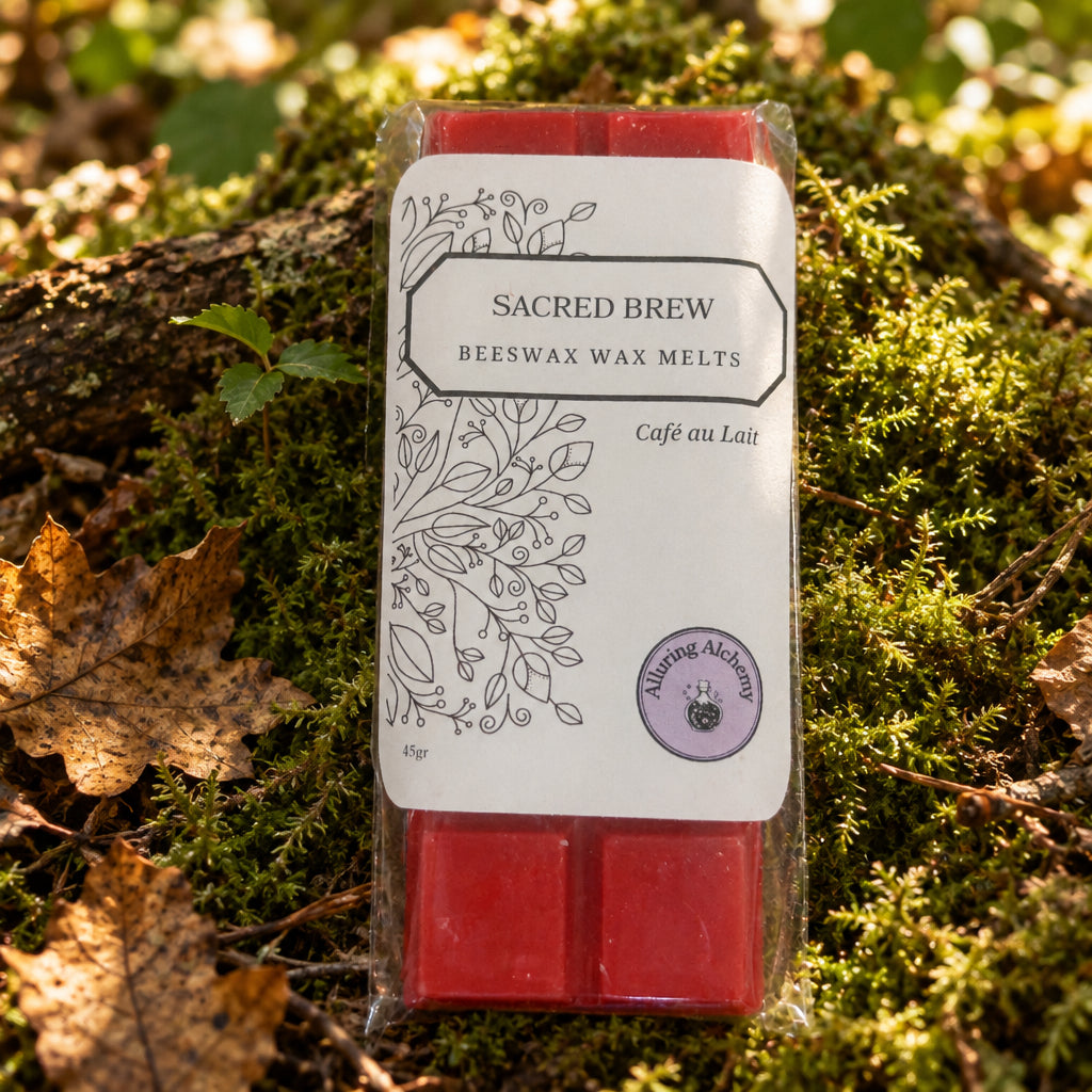 Sacred Brew Beeswax Wax Melts in a clear plastic package with a white label, placed on a light wood surface. The wax melts visible at the top and bottom are red, and the label reads 'SACRED BREW' and 'BEESWAX WAX MELTS' with 'Café au Lait' in script.
