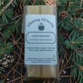 A packaged bar of Alluring Alchemy Ruby Reverie beeswax melts, placed on a forest floor surrounded by pine needles and pinecones.