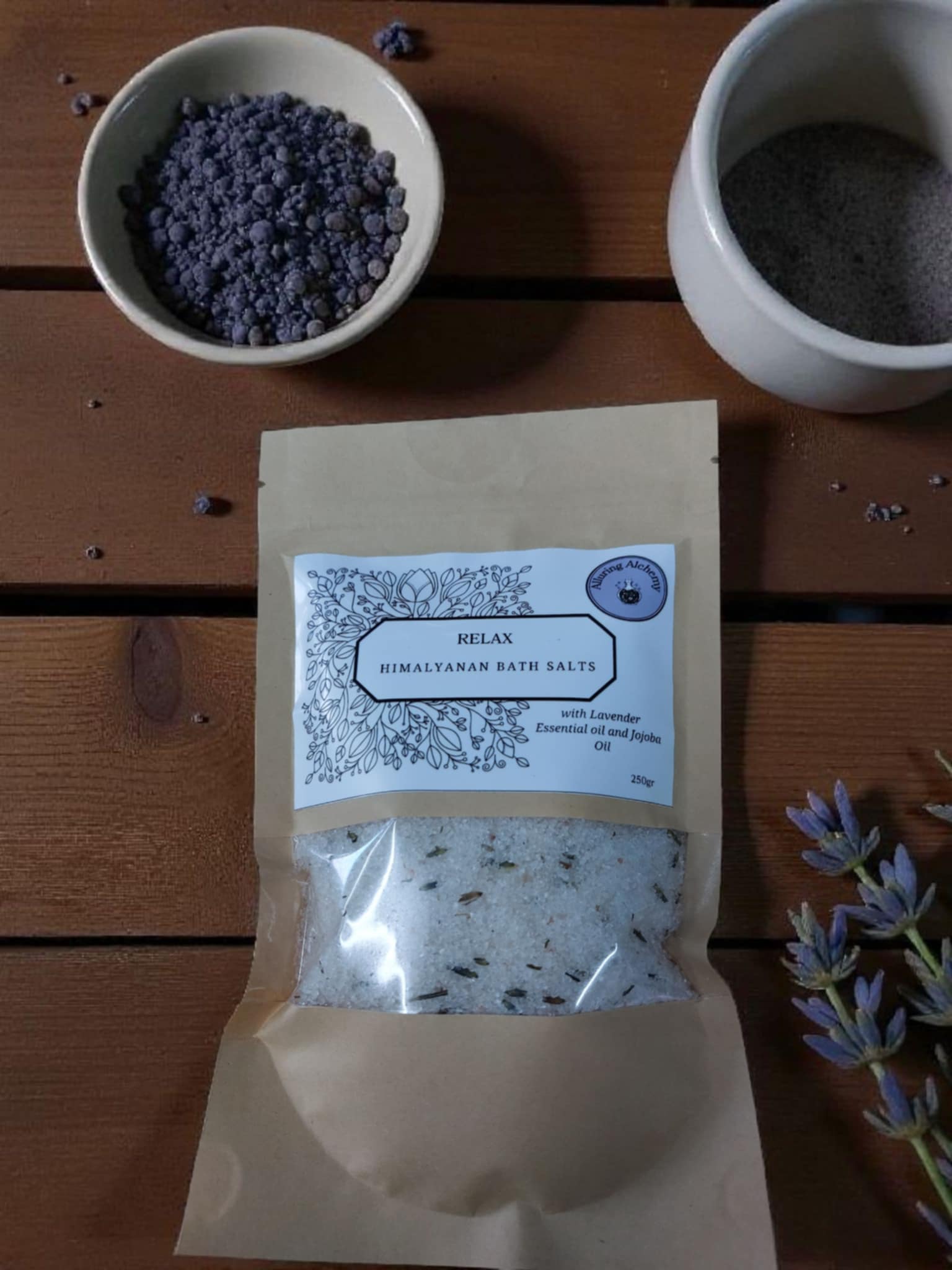 Packaged Himalayan bath salts with two bowls of purple and gray substances on a wooden surface.