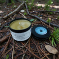 Candle in a jar labeled 'Earthwood' on a natural setting with pine needles and branches.