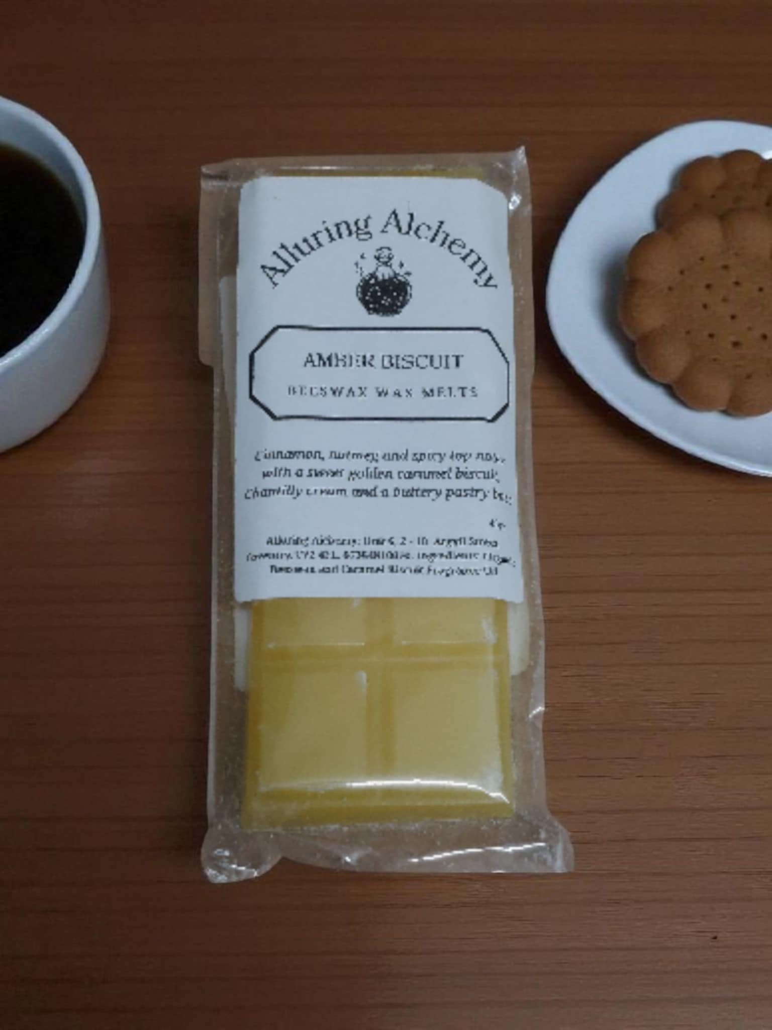 Packaged amber biscuit with a label on a wooden surface next to a cup of coffee and a plate of cookies.
