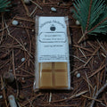 Wax melt package labeled 'Alluring Alchemy' on a natural background with pine needles and twigs.
