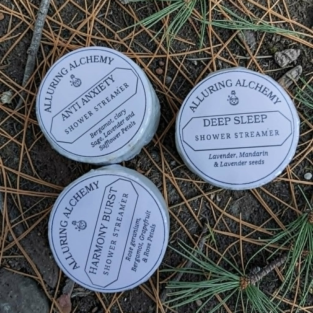 Three Alluring Alchemy shower streamer containers on a natural background with pine needles and branches.