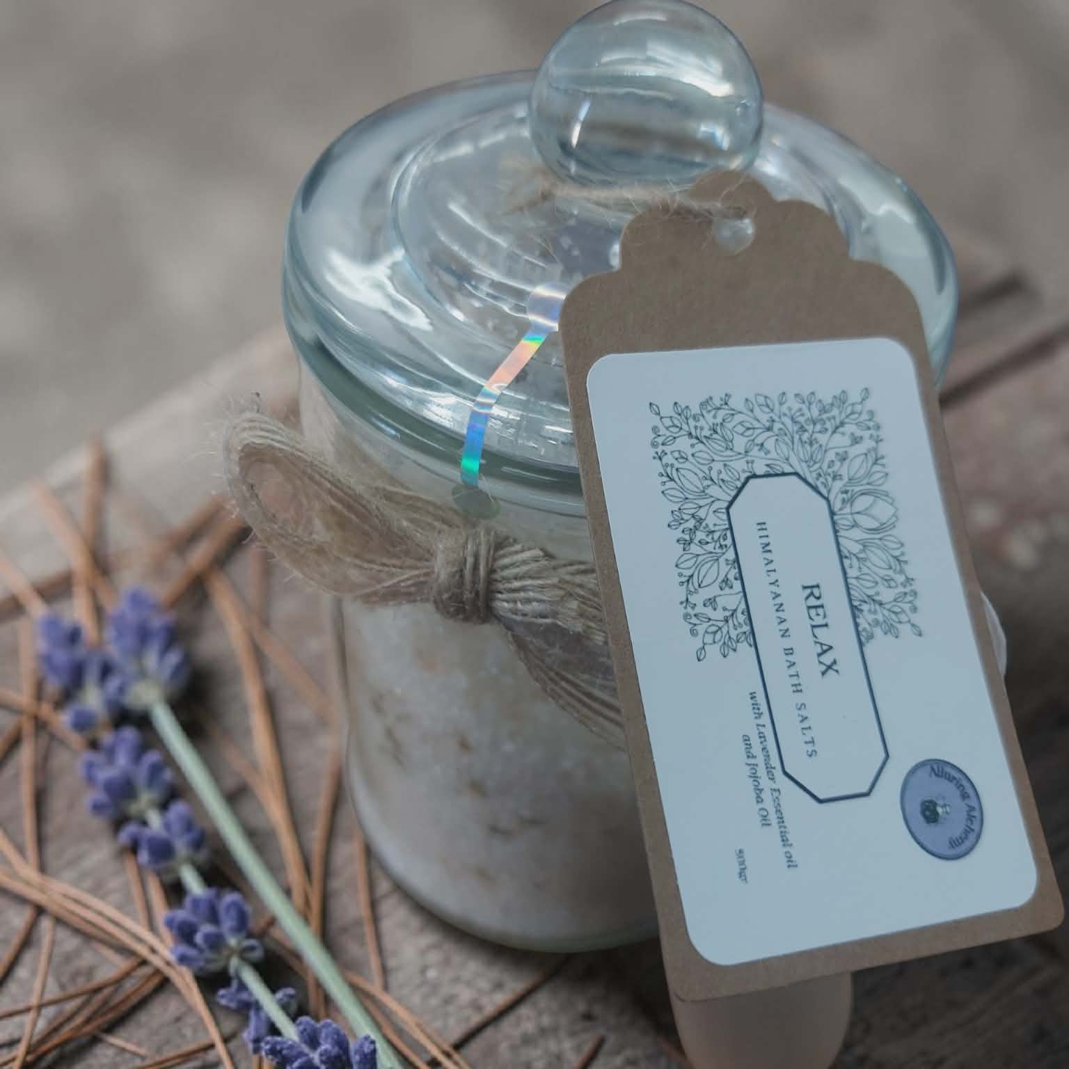 Jar of bath salts with a 'Relax' tag on a wooden surface with lavender and pine needles.