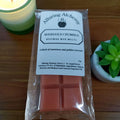 Pack of Alluring Alchemy rose gold crumble wax melts on a wooden surface with a candle and plant in the background.