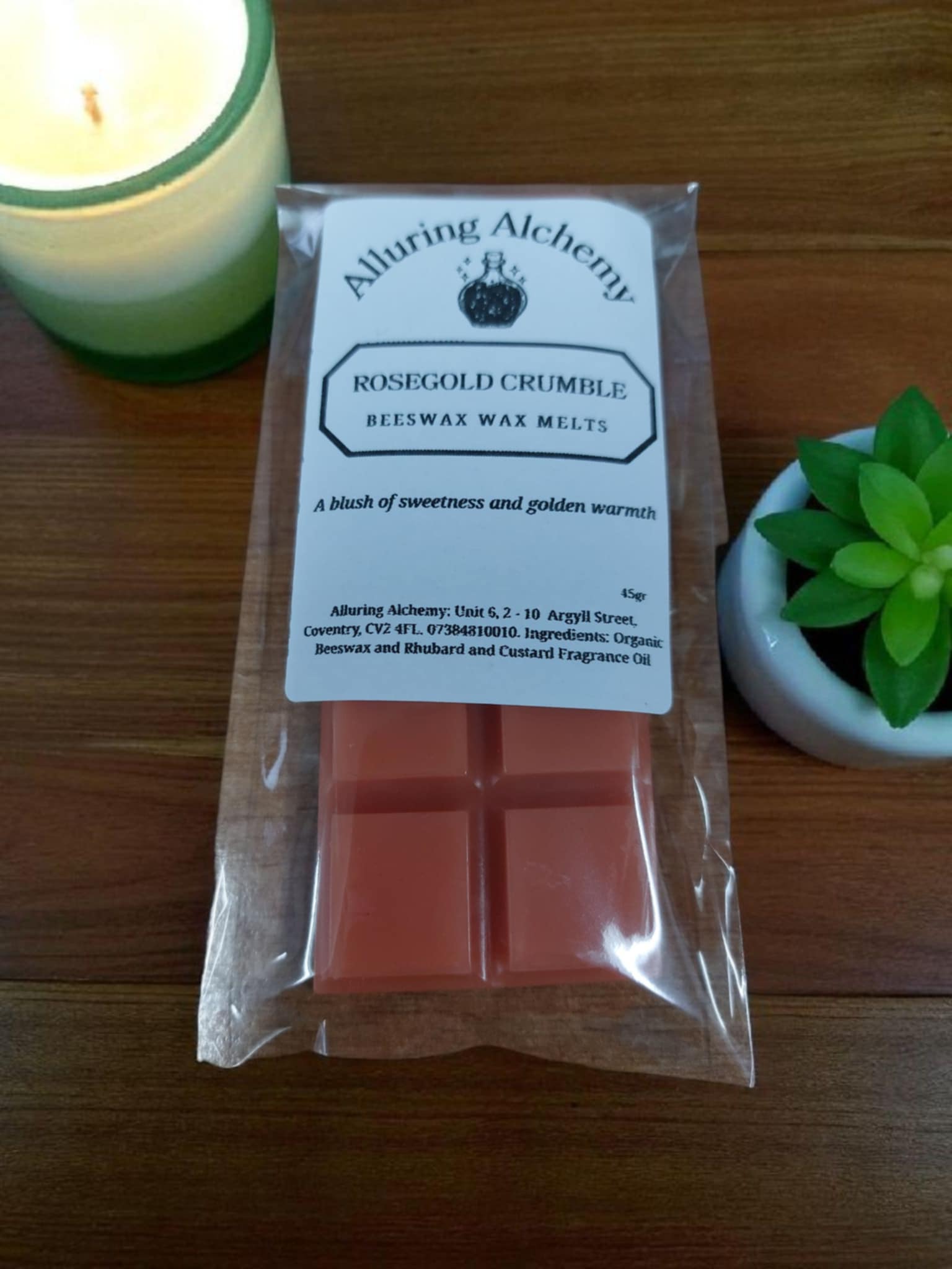 Pack of Alluring Alchemy rose gold crumble wax melts on a wooden surface with a candle and plant in the background.