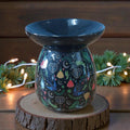 Decorative oil burner with colorful designs on a wooden stand, surrounded by festive lights and greenery.
