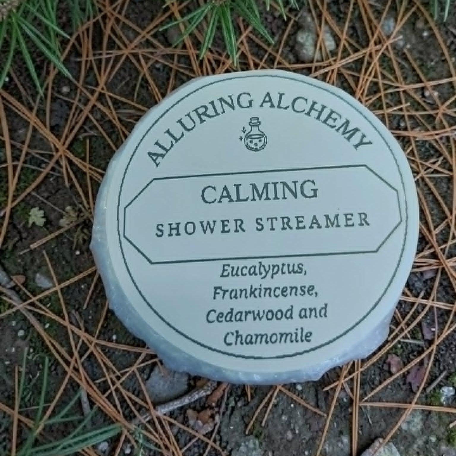Alluring Alchemy Calming Shower Streamer container on a natural background with pine needles and branches.