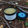 Candle in a black container with a label on a natural background of twigs and leaves.