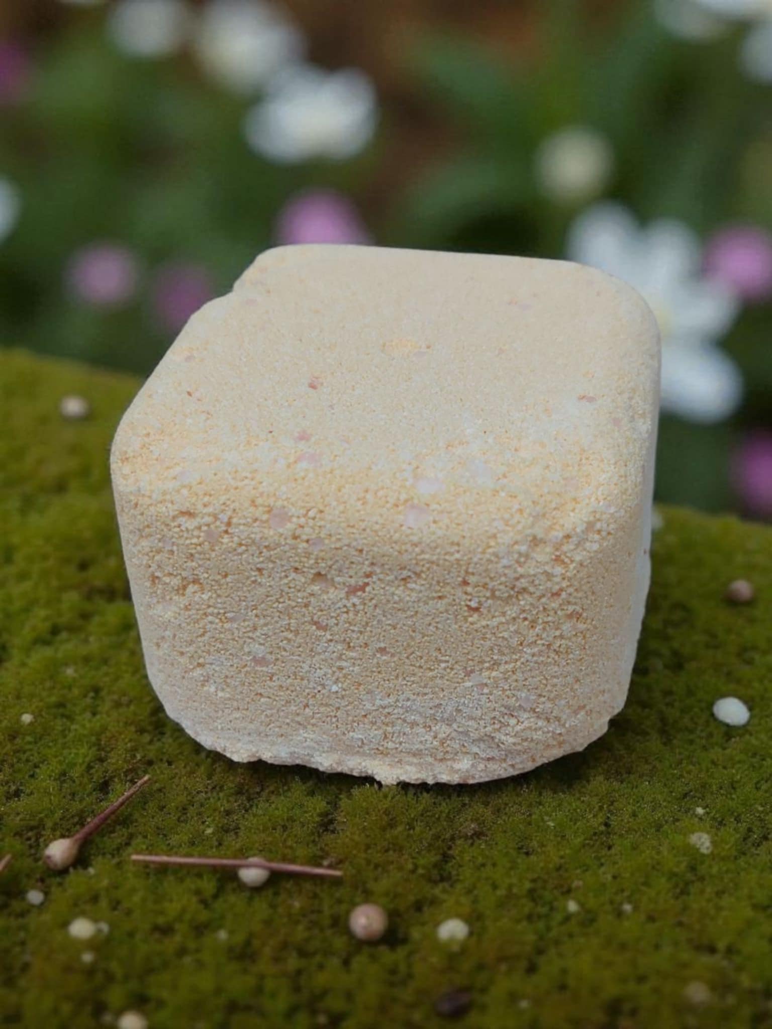 Square bath bomb on a green surface with blurred flowers in the background