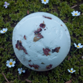 White bath bomb with natural elements on a green moss background with small white flowers