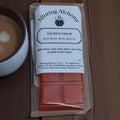 Pack of Alluring Alchemy Sacred Brew beeswax wax melts on a wooden surface with a cup of coffee.