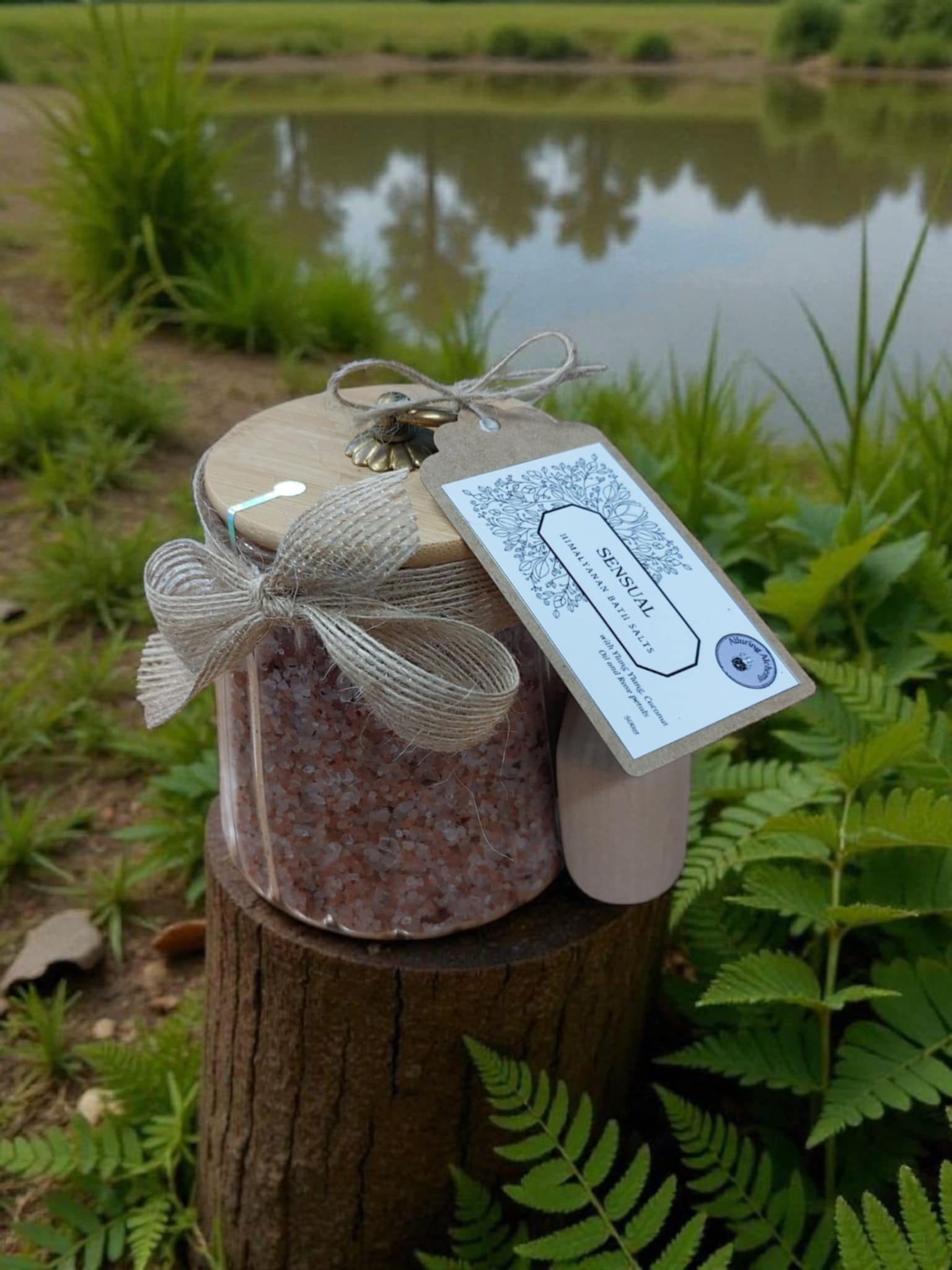 Jar of pink bath salts with a decorative bow and tag on a wooden stump by a pond.
